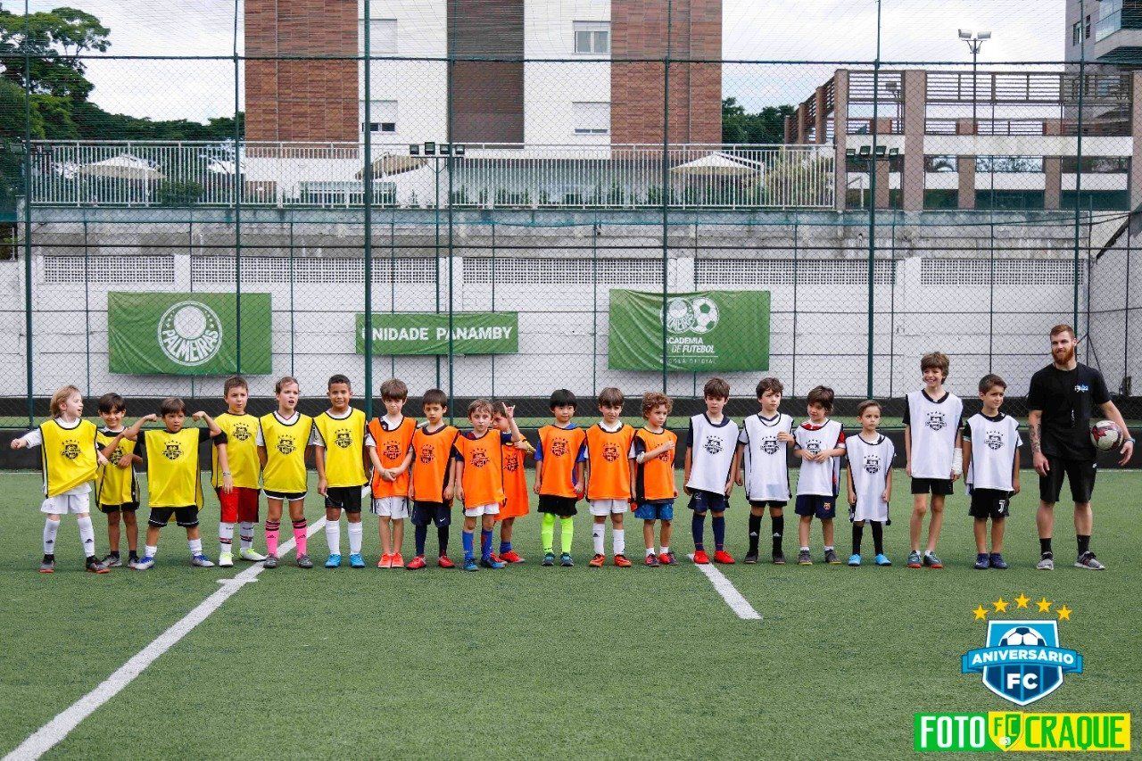 Crianças com uniformes de futebol alinhadas em um campo verde com um treinador, faixas ao fundo.