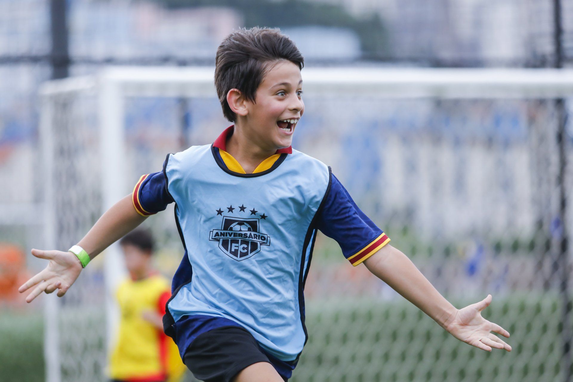 Menino comemora no campo de futebol com os braços estendidos, vestindo uma camisa azul e sorrindo.