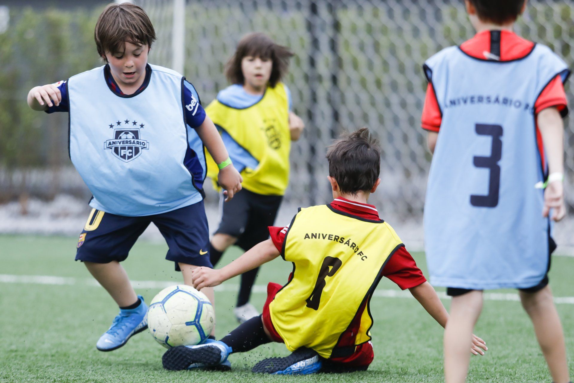 Crianças jogando futebol em um campo verde; uma delas está caindo, enquanto outras usam coletes numerados com 1, 3 e sem identificação.