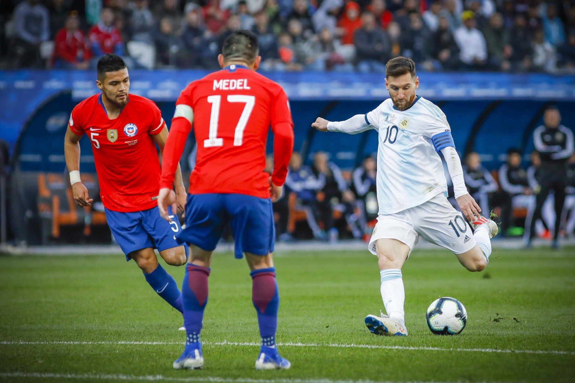 Lionel Messi chutando uma bola de futebol em um jogo contra o Chile. Jogadores com uniformes vermelhos e azuis no estádio.