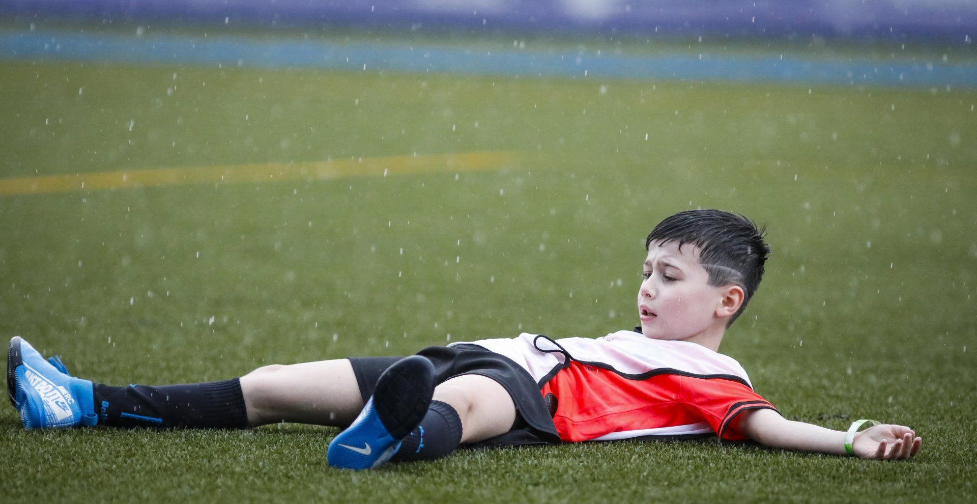 Menino com camisa de futebol vermelha deitado em um campo molhado, com aparência cansada sob a chuva.