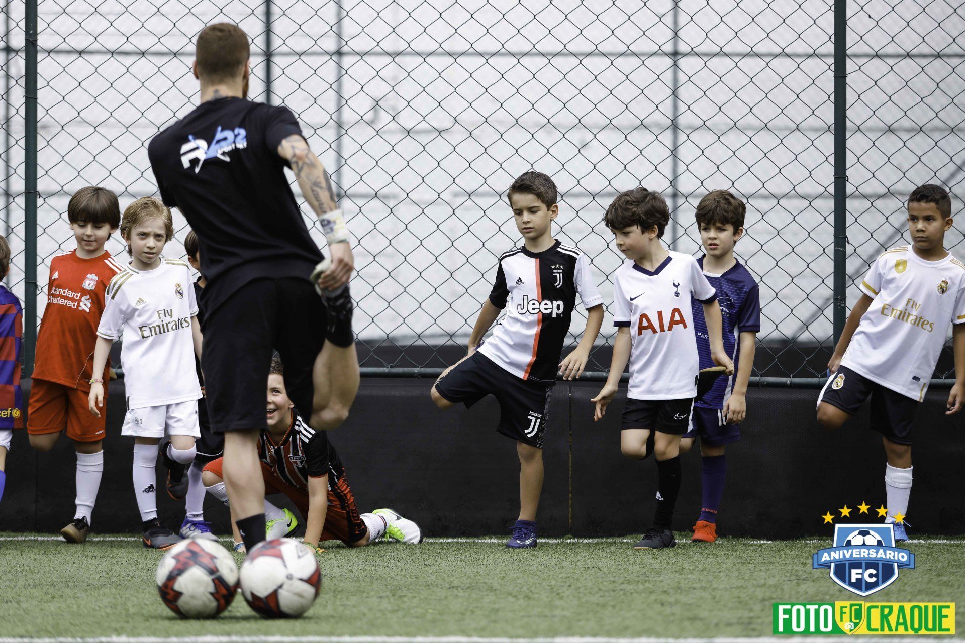 Treinador de futebol demonstrando um alongamento para um grupo de jovens jogadores em um campo de grama sintética.
