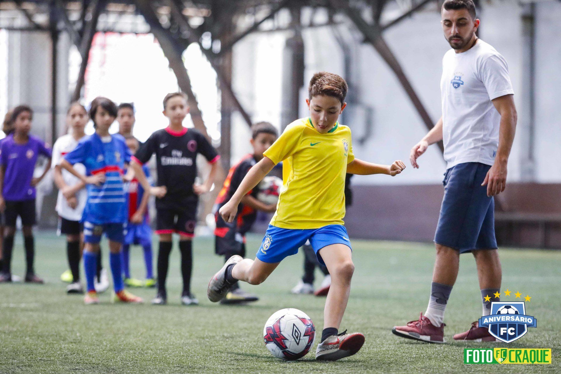 Menino de camisa amarela chutando uma bola de futebol em quadra coberta, outras crianças assistindo, treinador por perto.