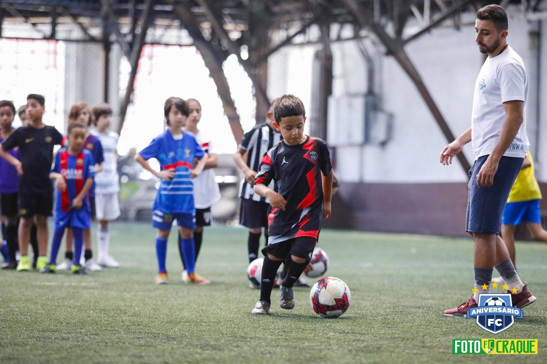 Crianças com uniformes de futebol praticam exercícios com um treinador em um campo coberto.