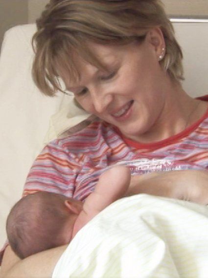 Eine frisch gebackene Mutter legt ihr Baby an die Brust und schaut mit einem warmen Lächeln -aus dem Film.