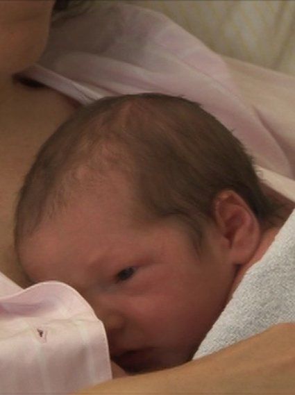 Ein waches Neugeborenes liegt Haut an Haut auf der Brust seiner Mutter, in ein weiches Tuch gehüllt.
