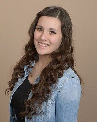 Woman with long brown curly hair smiles, wearing a black shirt and denim jacket, against a neutral background.