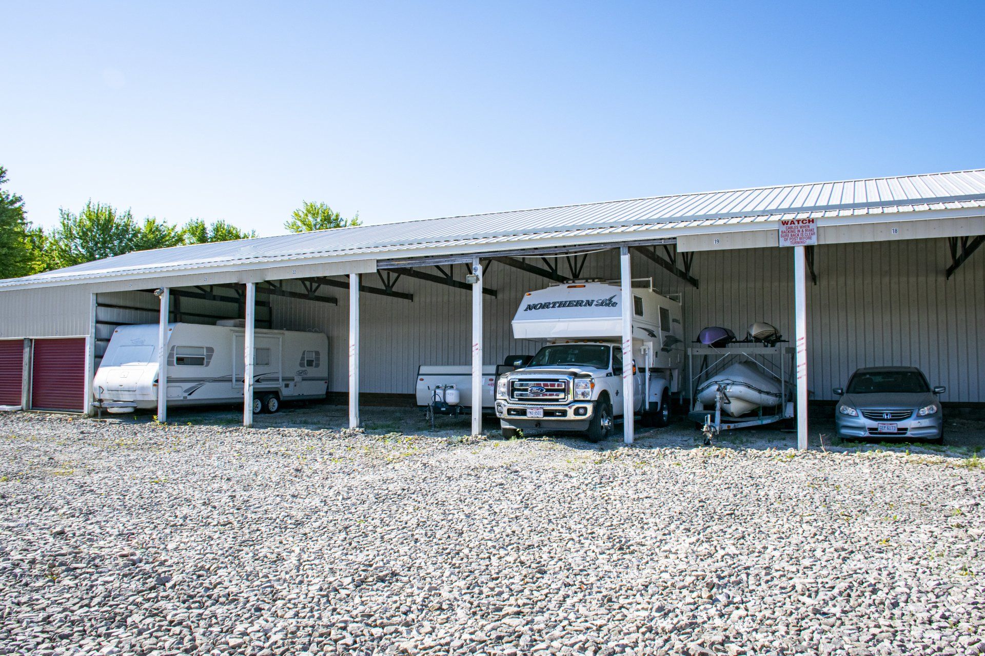 Covered RV and vehicle storage units; gravel ground, sunny day.