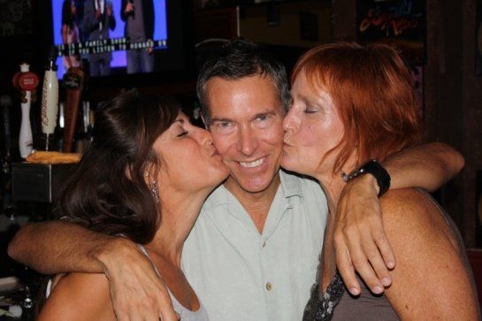 Two women kissing a man on the cheek in a bar