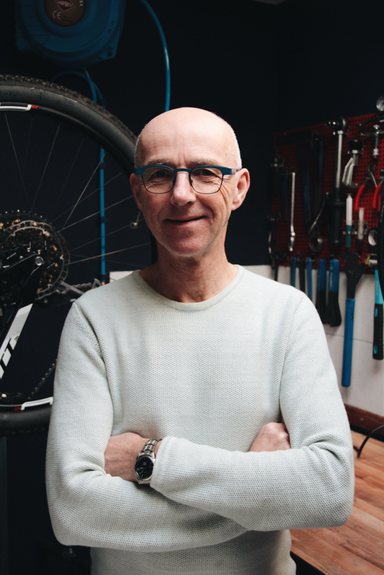 Een glimlachende kale man in een witte trui staat met de armen over elkaar in een fietsenwerkplaats.