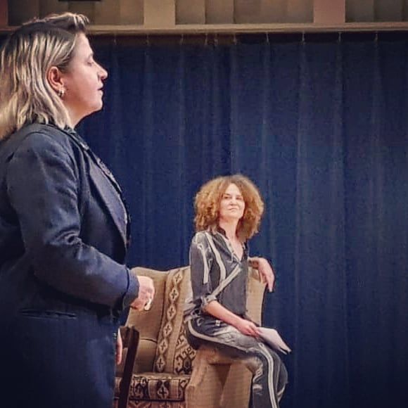 Twee vrouwen op een podium, de ene staand in profiel en de andere zittend op een stoel tegen een donker gordijn.