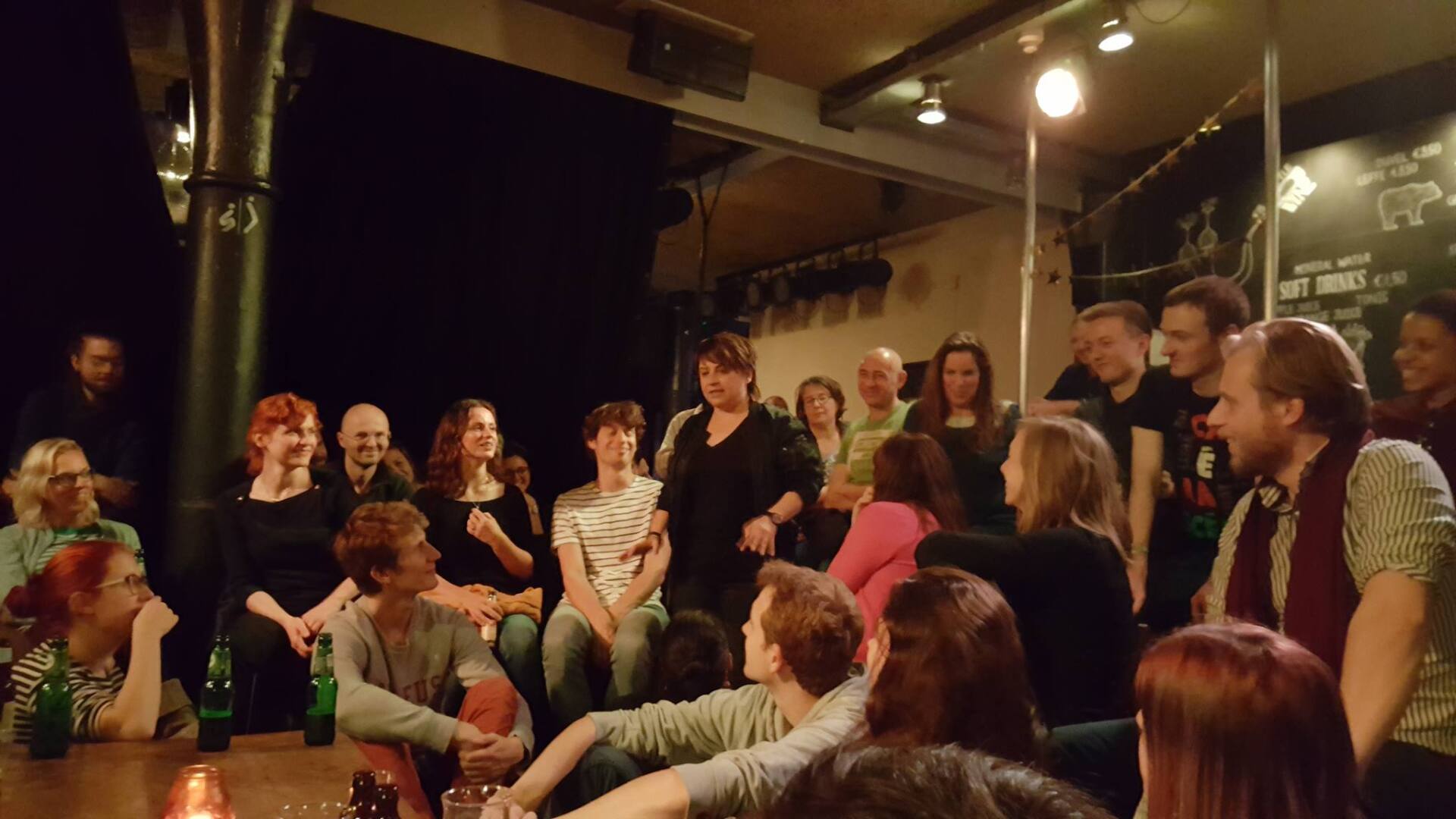 Het publiek was bijeengekomen in een sfeervol verlichte ruimte, zittend en staand rond een spreker tijdens een klein evenement.
