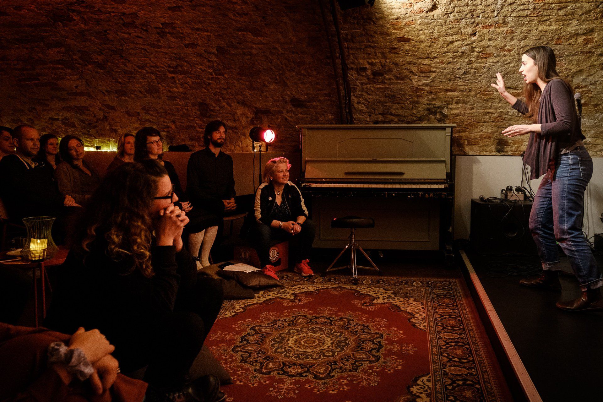 Het publiek kijkt naar een spreker op een klein podium in een warm verlichte ruimte met een piano en een tapijt met een patroon.