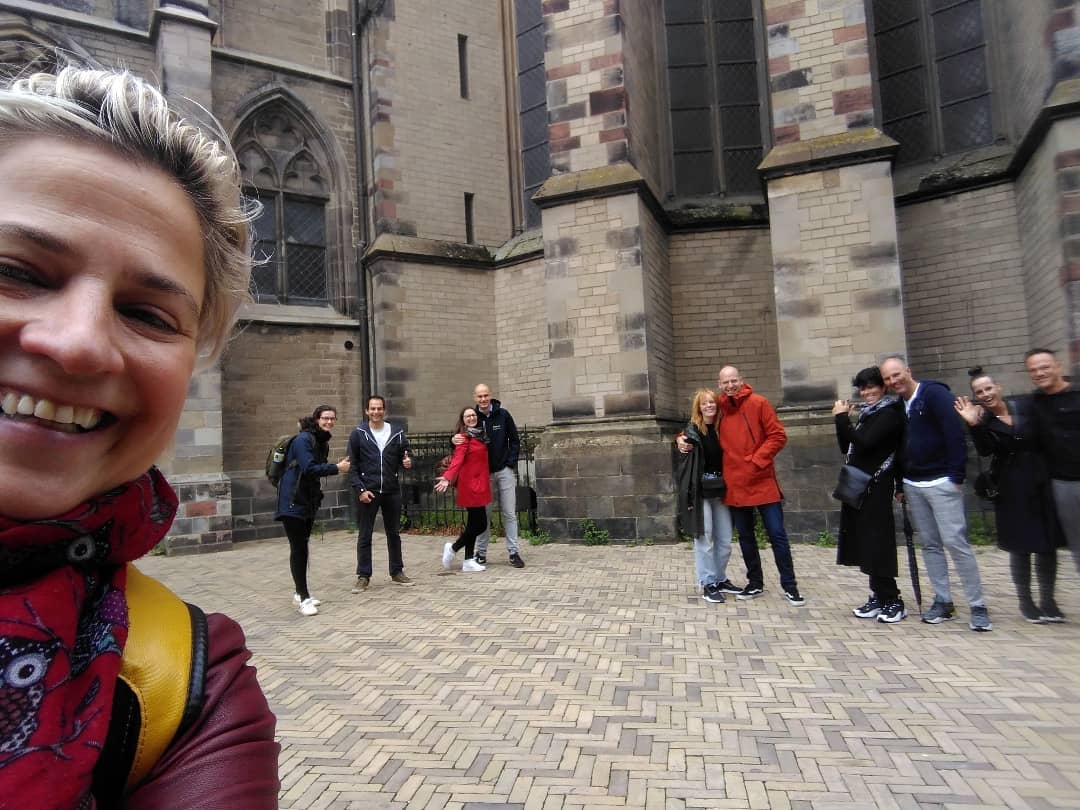 Een lachende vrouw op de voorgrond maakt een selfie met een groep mensen buiten een stenen kathedraal.