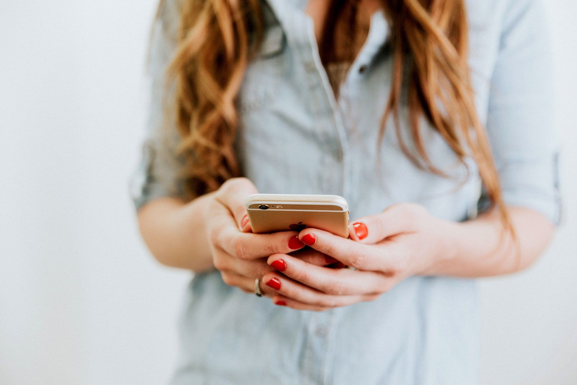 Person holding a smartphone with both hands, wearing a light blue shirt with red painted nails