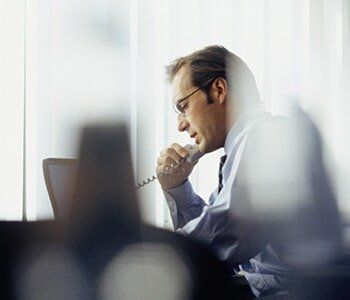 Man in glasses talking on a landline phone, looking at a laptop in an office.