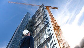 Construction worker in hardhat looking up at a tall building under construction with a crane, sunny sky.