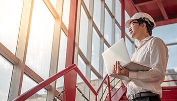 Engineer in hardhat, holding laptop, looking out a window. Red stairs, bright sunlight.