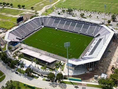 Aerial view of a football stadium with green field, grey seating, and parking lots.