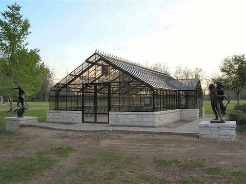 Greenhouse with glass walls, black metal frame, and sculptures on either side in a park.