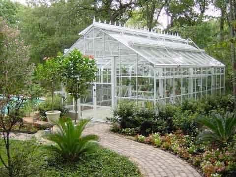 White greenhouse with glass walls and roof, surrounded by lush greenery and a brick path.