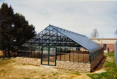 Greenhouse with glass roof and walls, set on a stone foundation, near a tree and building.