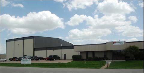 Exterior view of a light-colored industrial building under a partly cloudy sky; cars parked in front.