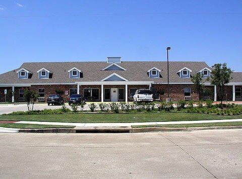 Brick building with a gray roof, dormers, and a lawn. Cars are parked in front.