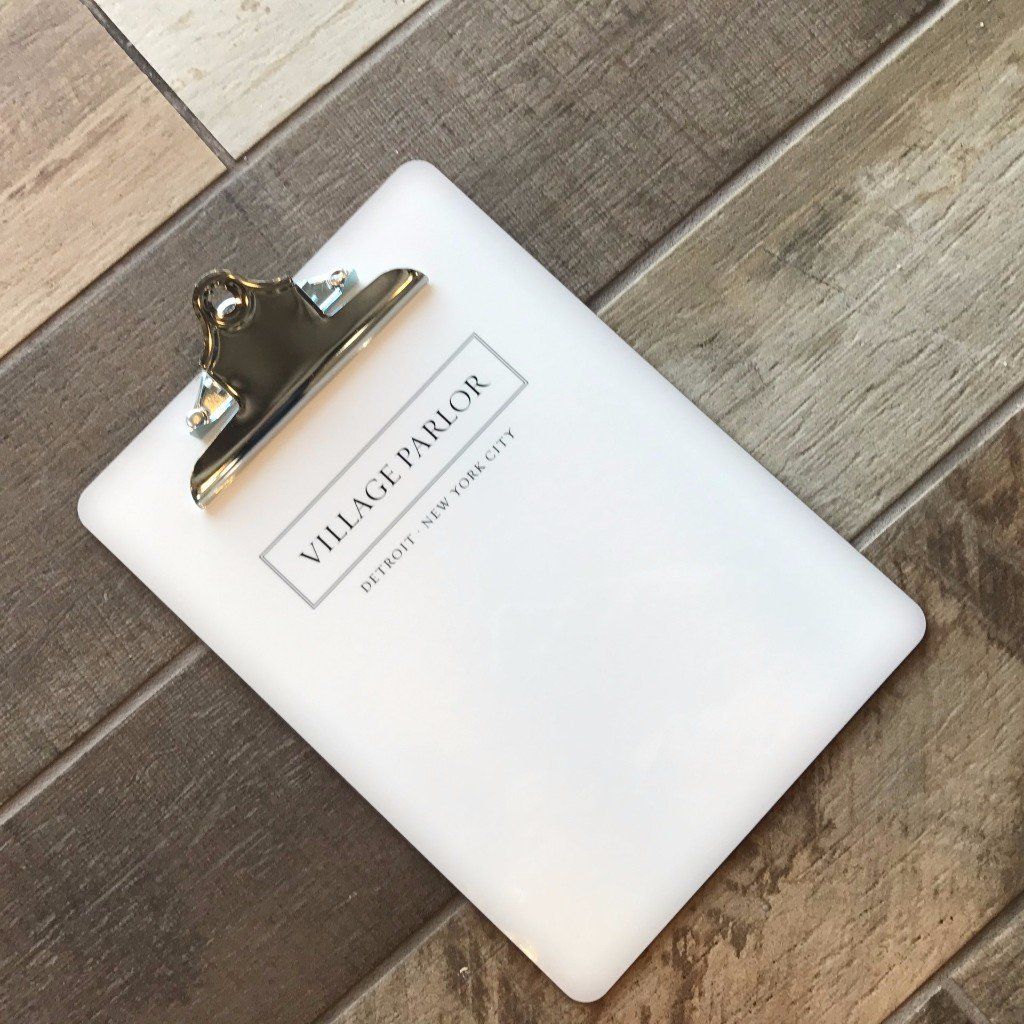 White clipboard with “Village Parlor” label on a rustic wood floor.
