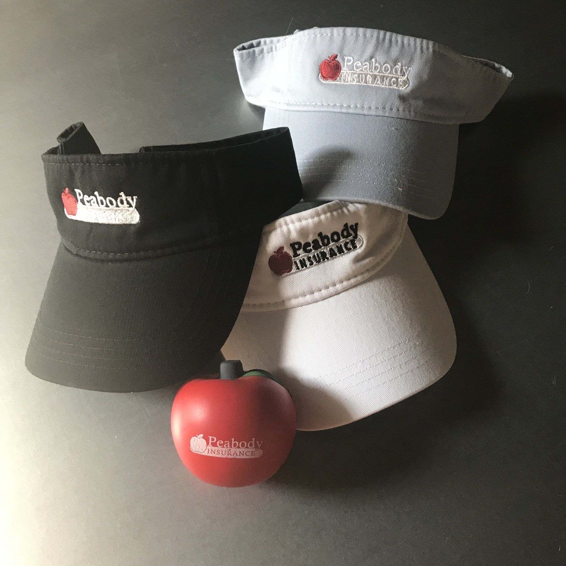 Three Pebody-branded visors in black, white, and gray with a red apple on a dark surface.