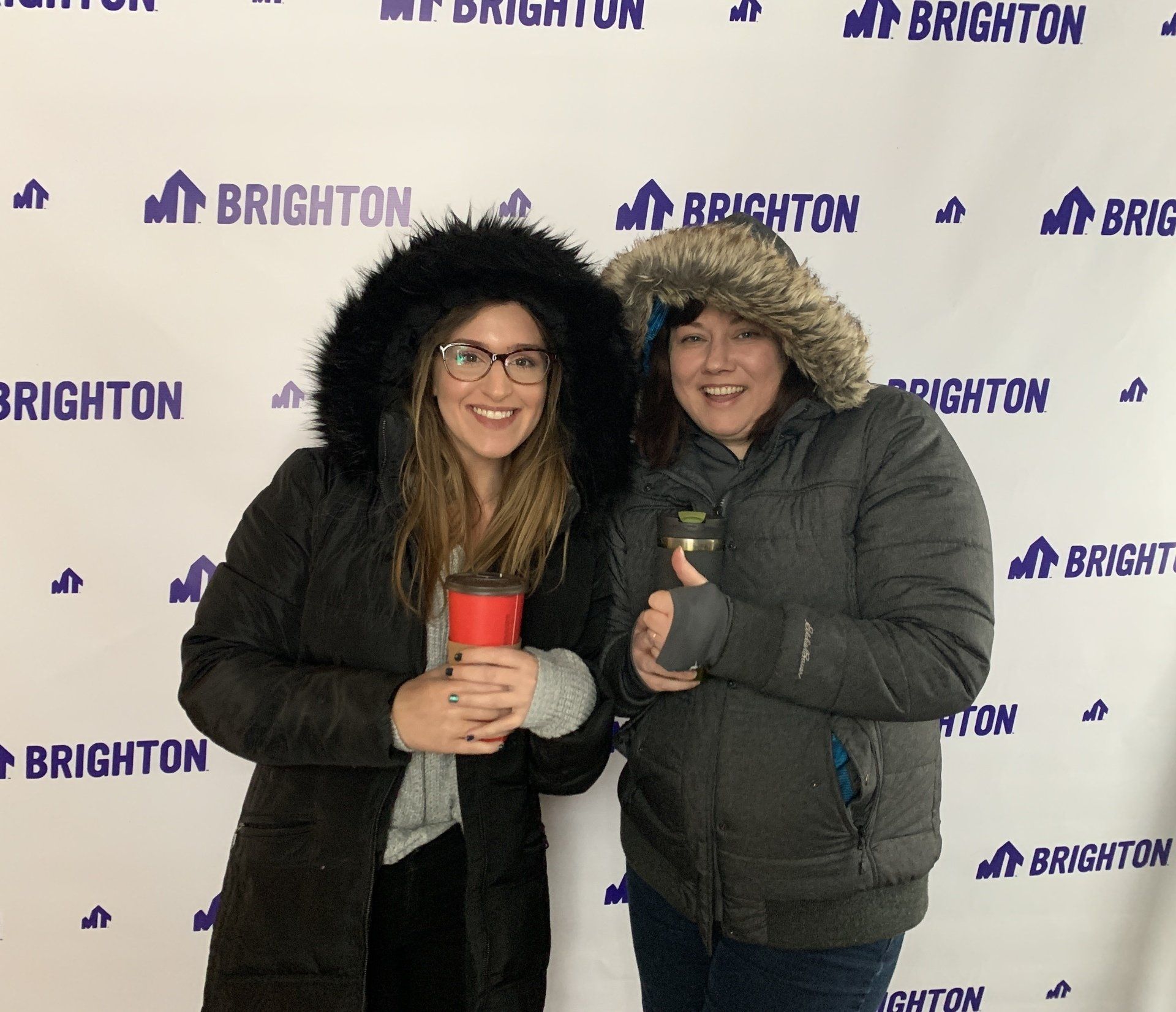 Two people smiling at a Brighton event backdrop, holding cups and wearing winter coats and hats.