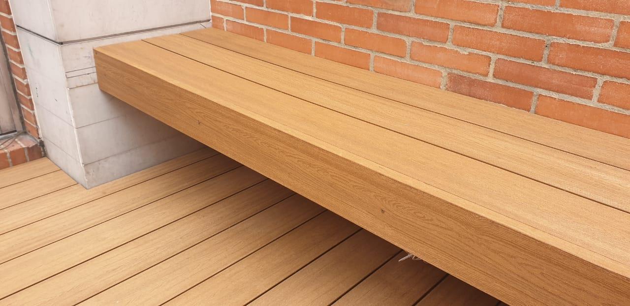 un banco de madera está sobre una terraza de madera junto a una pared de ladrillos.