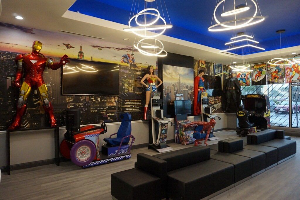 Playroom with superhero statues, TV, arcade machines, and black seating under blue ceiling lights