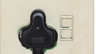 Black electrical plug inserted into outlet, next to a double light switch, on a beige wall.