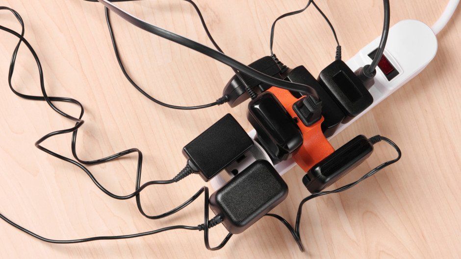 Power strip overloaded with black chargers, tangled cords on wood surface.