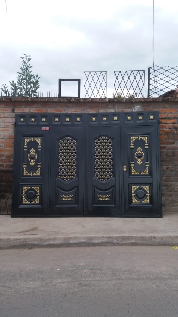 Una puerta de metal negro de doble panel con detalles ornamentales dorados, situada frente a un muro de ladrillo