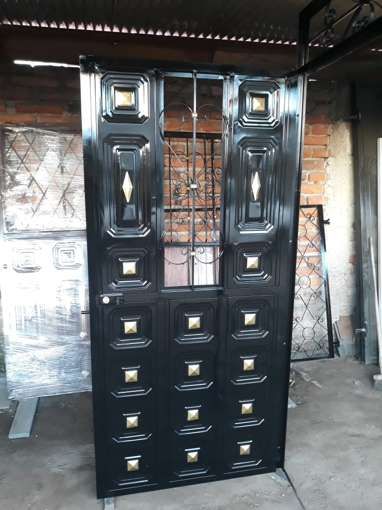 Puerta de metal negro con una ventana central decorativa con barrotes y detalles en color dorado, ubicada en un taller.