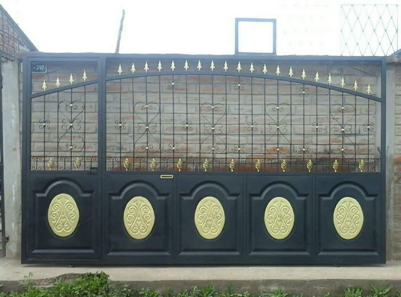 Puerta de metal negro con paneles ovalados dorados y remates decorativos, colocada contra una pared de ladrillo.
