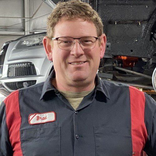 Smiling mechanic in a red-trimmed work shirt standing in a garage with a car hood open