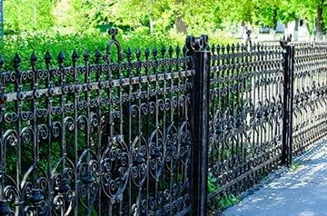 Black wrought-iron fence along a sunlit sidewalk with green trees in the background