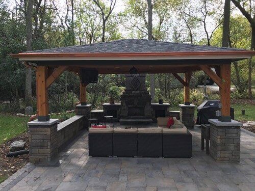 Covered outdoor pavilion with stone fireplace and seating in a wooded area