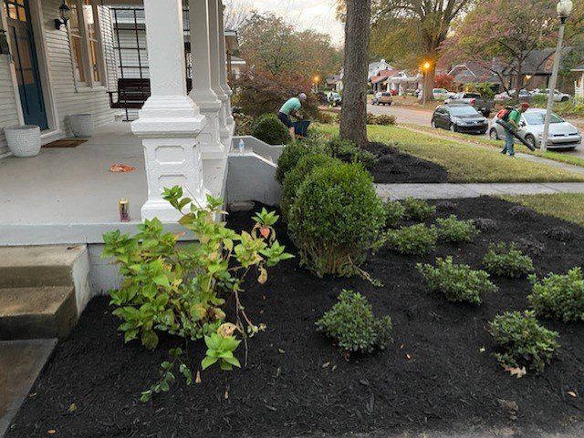 Workers landscaping a front yard with fresh mulch; shrubs by a porch, street in the background.