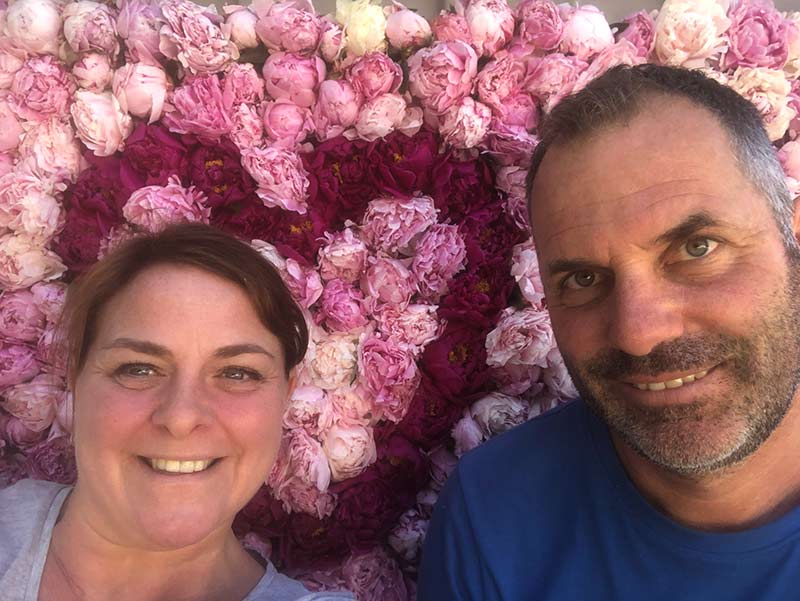 Couple souriant devant un mur composé de pivoines roses et bordeaux