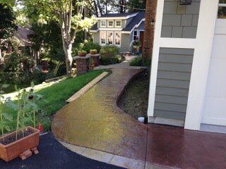 Stamped concrete walkway curves toward a light green house with gray siding; a lawn and driveway border the path.