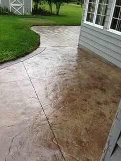Stamped concrete patio, brown and tan, next to a light gray house and green grass.