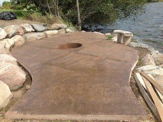 Concrete patio with fire pit by a lake, surrounded by rocks.