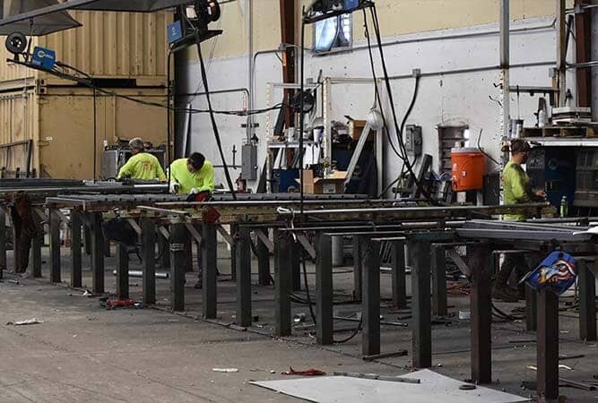 Workers in a metal fabrication shop are cutting and assembling metal pieces at workbenches.