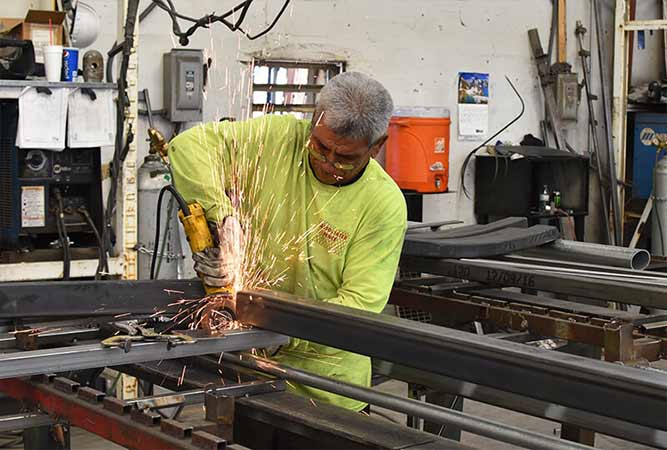 Man in yellow shirt grinding metal, creating sparks, inside a workshop.