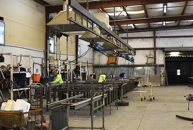 Workshop interior with workers welding metal on long workbenches under overhead exhaust hoods.