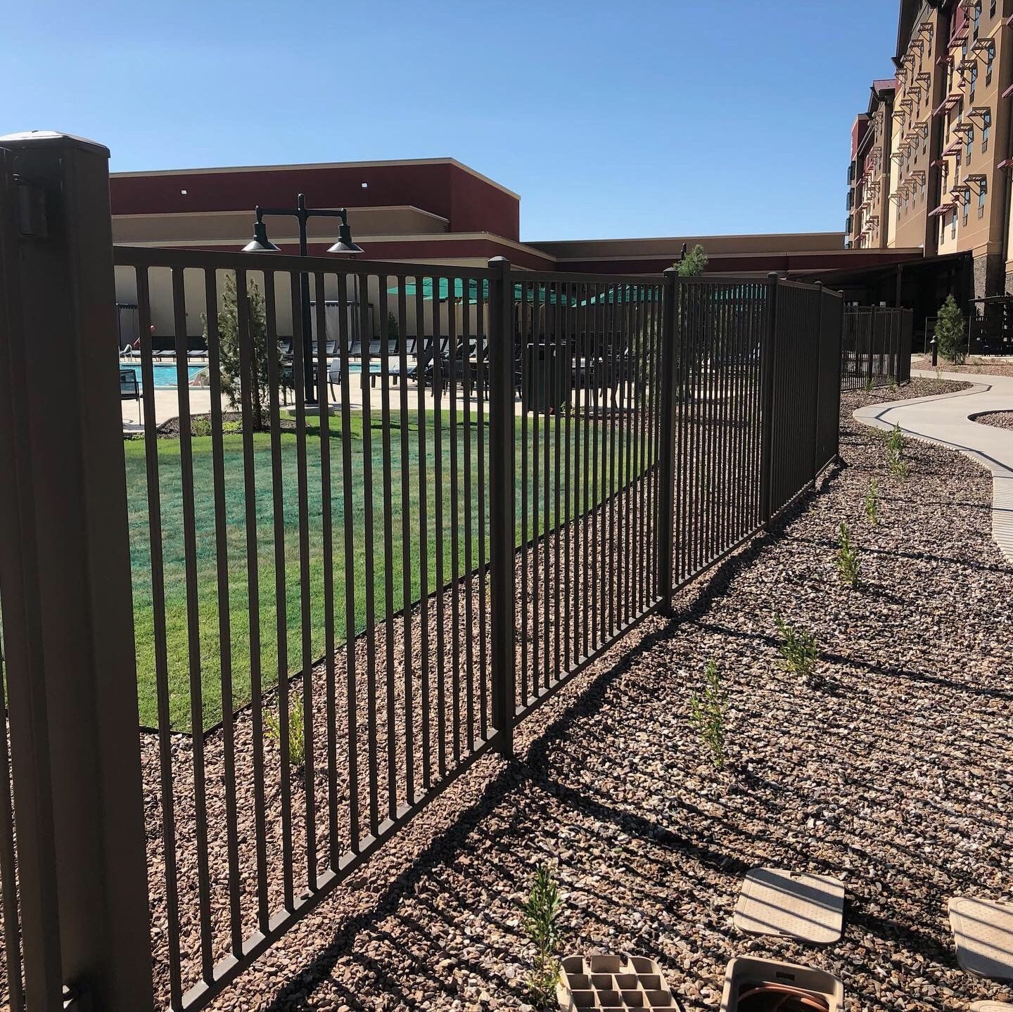 Brown metal fence surrounding a grassy area next to a path and building on a sunny day.